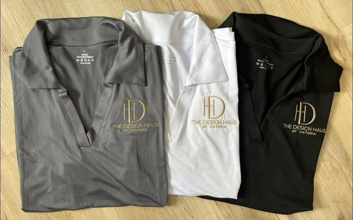Three folded polo shirts in grey, white, and black with gold embroidered logo and text that reads "The Design Haus of Victoria"