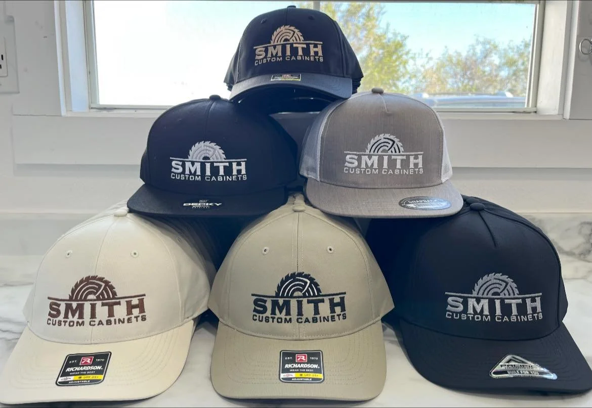 Six baseball caps in various colors with the logo and text 'SMITH CUSTOM CABINETS' on the front, arranged in a pyramid shape on a white surface with a window in the background.