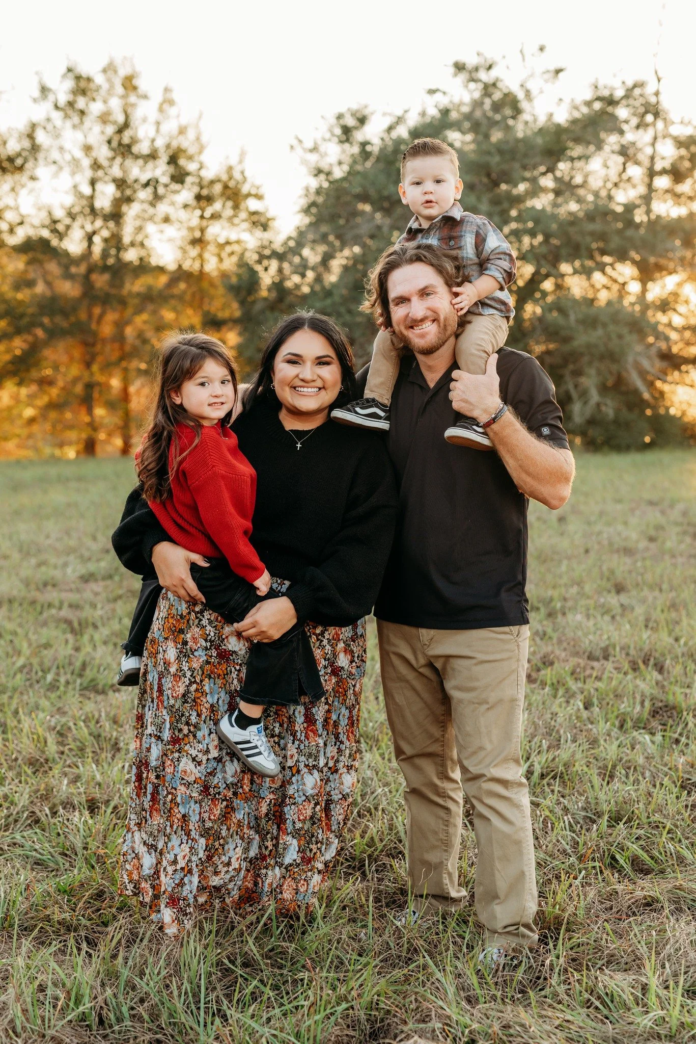 A family of four standing in a grassy field during sunset, smiling at the camera. The mother is holding a young girl dressed in a red sweater, the father is carrying a young boy on his shoulders, and the background features trees with autumn foliage.