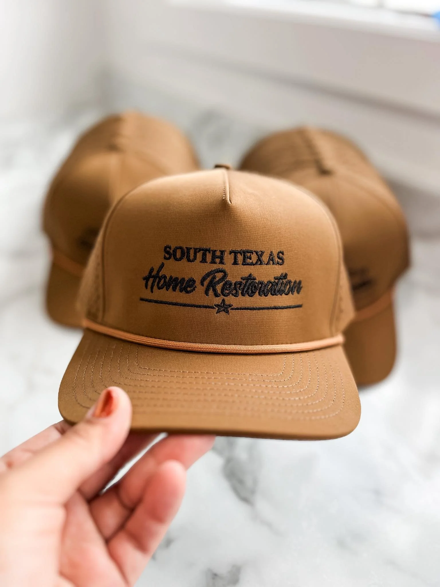A tan baseball cap with black embroidered text reading 'South Texas Home Restoration,' held by a person's hand against a white marble background with three more tan caps in the background.