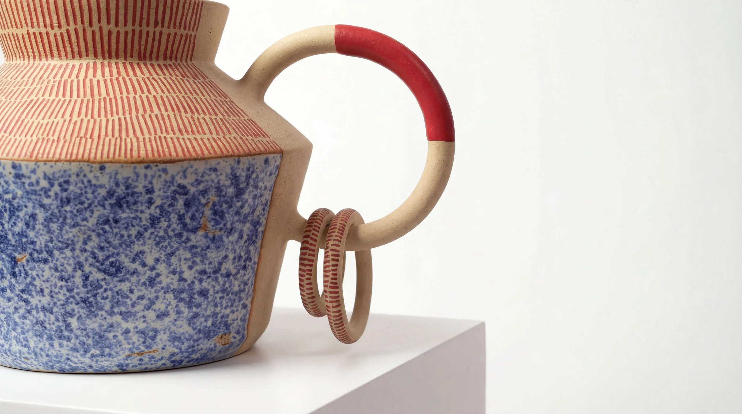 Close-up of a colorful ceramic pitcher with a textured blue lower half, red striped horizontal patterns, a rounded handle with a red top section, and a matching set of three red striped bangles hanging on the handle, positioned on a white surface aga