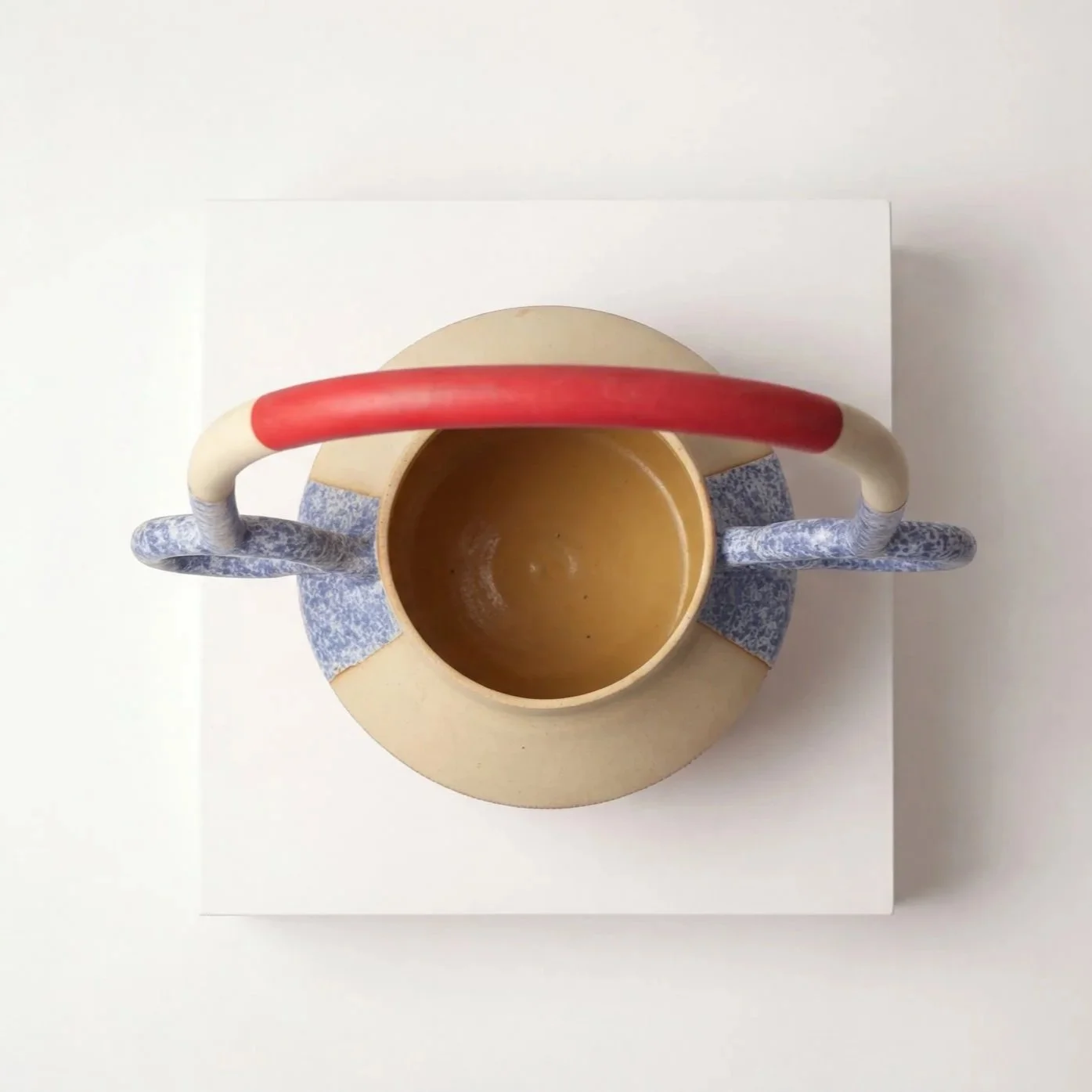 Top view of a round beige ceramic teapot with a red and beige handle, sitting on a blue and white patterned trivet on a white surface.