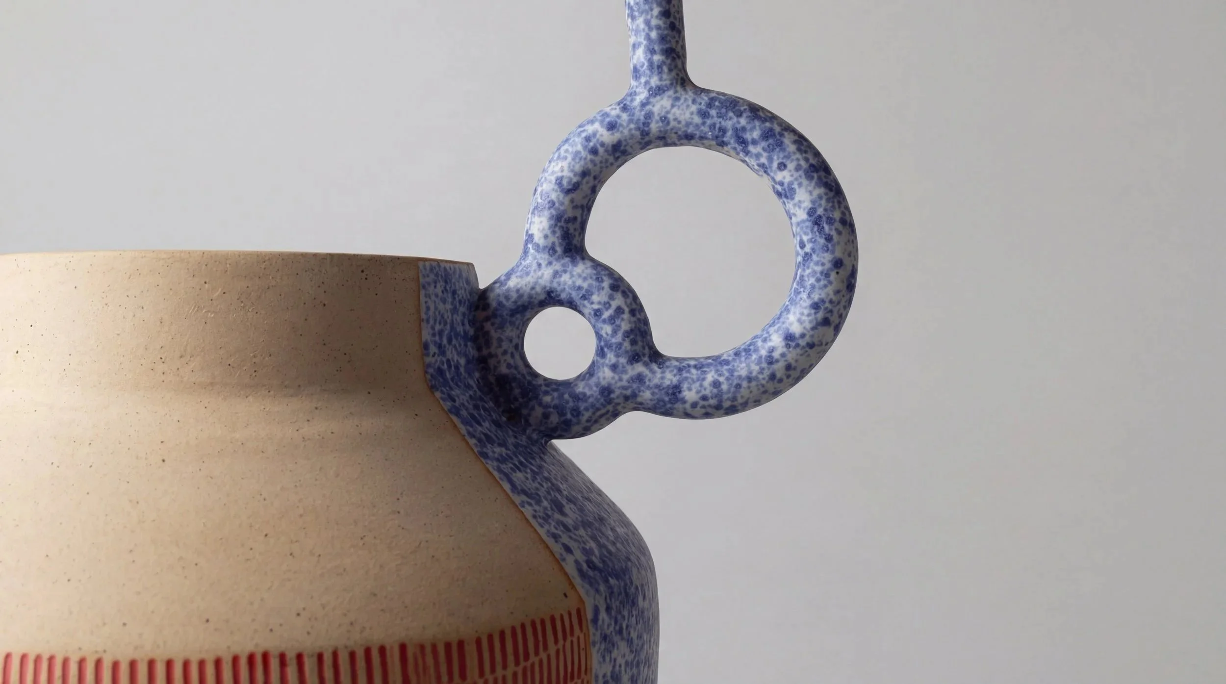 Close-up of a ceramic pitcher with a rough beige body and a speckled blue handle and spout, set against a plain gray background.