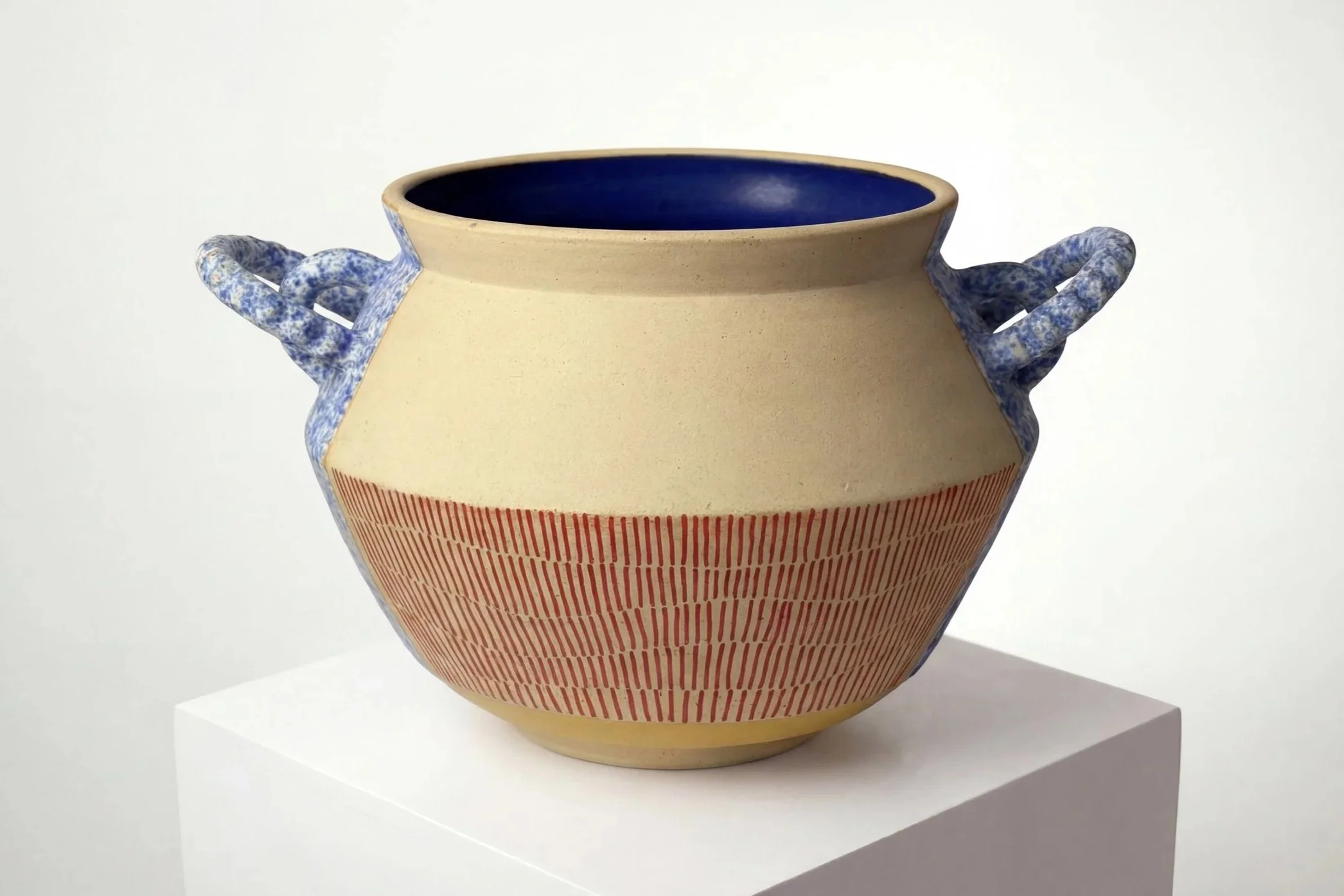 Ceramic pot with blue handles and a red striped pattern on the lower half, displayed on a white pedestal against a plain background.