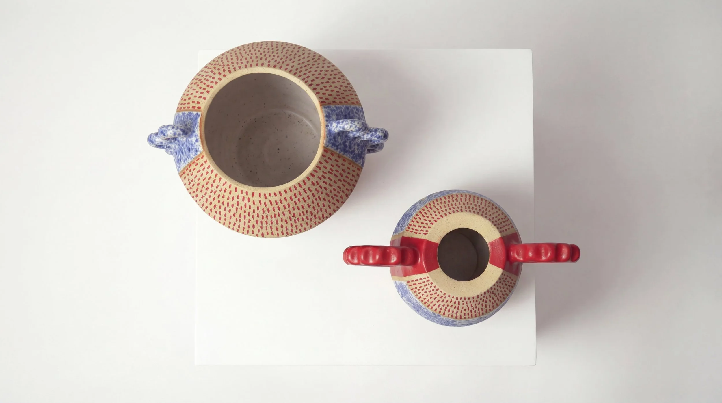 Top view of two decorative ceramic mugs with matching patterns, one with a red handle and the other with a blue handle, placed on a white surface.