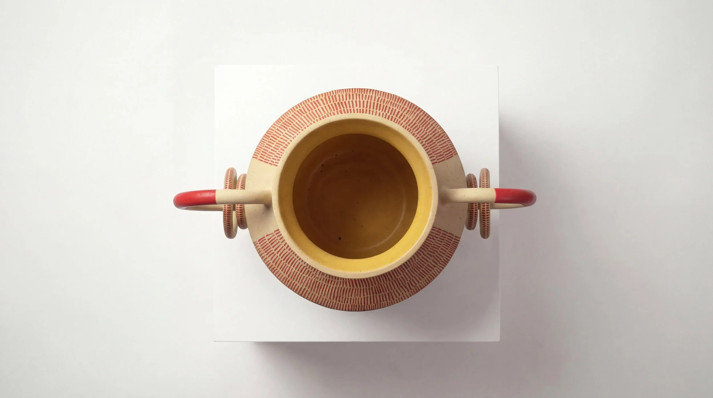 Top view of a decorative ceramic mug, with a red and beige pattern around the rim and a red handle, placed on a white square platform against a light-colored surface.