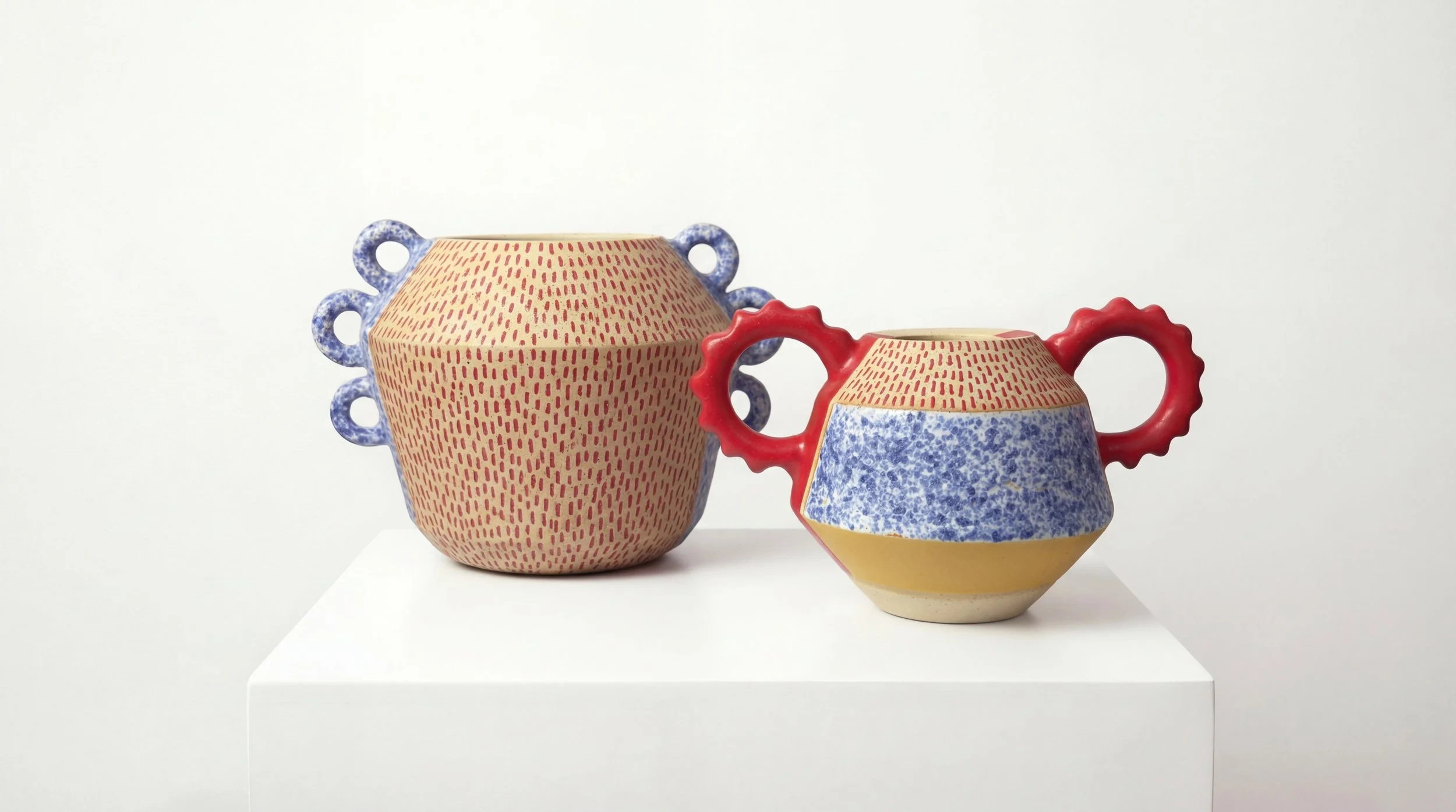 Two colorful ceramic vases with unique patterns and handles displayed on a white pedestal against a plain white background.