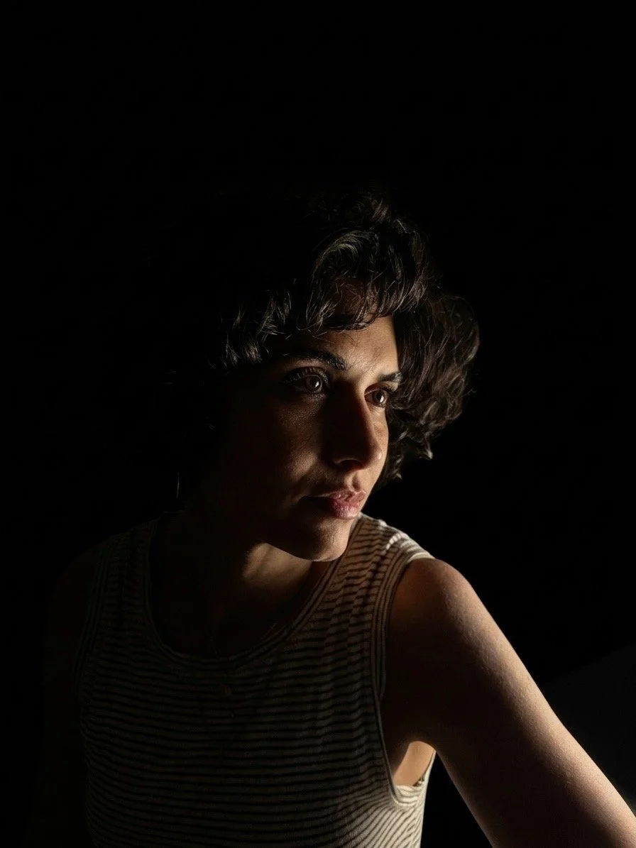 A woman with short curly hair looking to the side, lit from the front left, against a dark background.