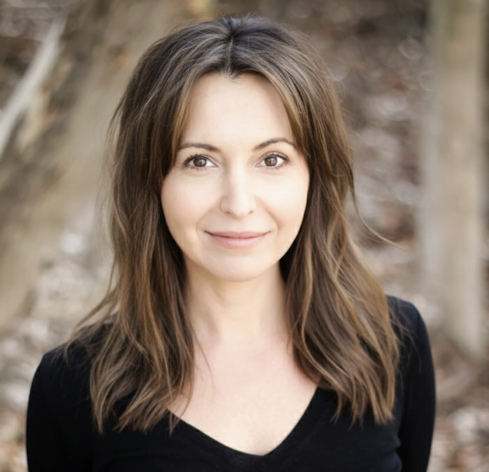 A woman with shoulder-length brown hair, smiling softly, standing outdoors with trees in the background.