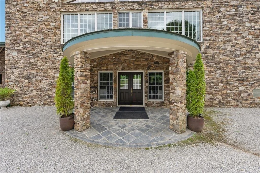 The image shows the entrance of a building with a stone facade, black double doors, and two large potted green trees flanking the door. There is a curved roof canopy over the entrance and a stone tiled porch area.