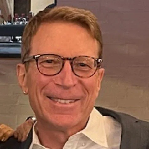 A man with short light brown hair, glasses, and earrings smiling at the camera. He's wearing a white shirt and a dark jacket. The background shows a wall and a bar area.