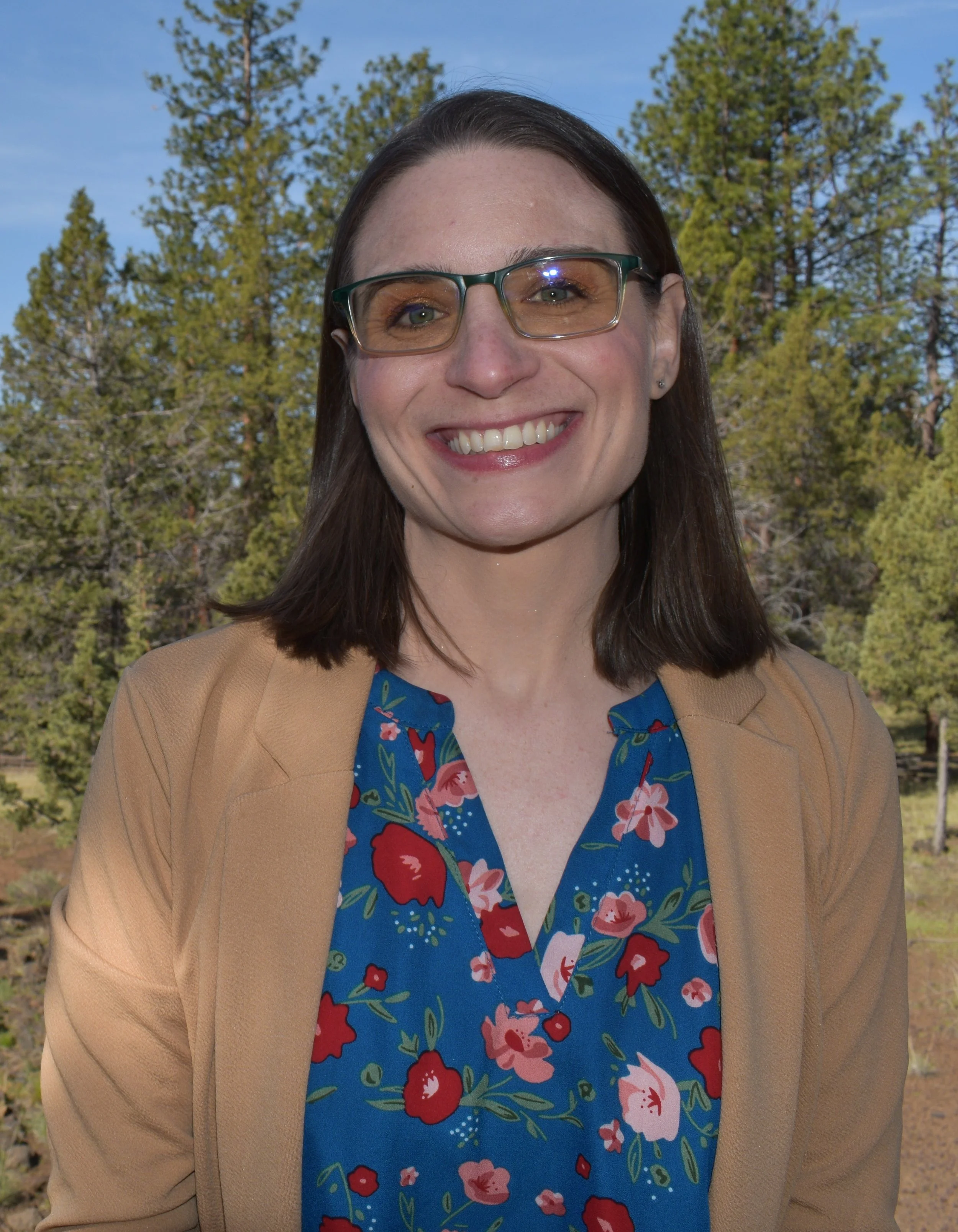 Close-up of a smiling woman with dark hair, eyeglasses, wearing a blue floral dress and tan blazer, standing outdoors with trees and a blue sky in the background.