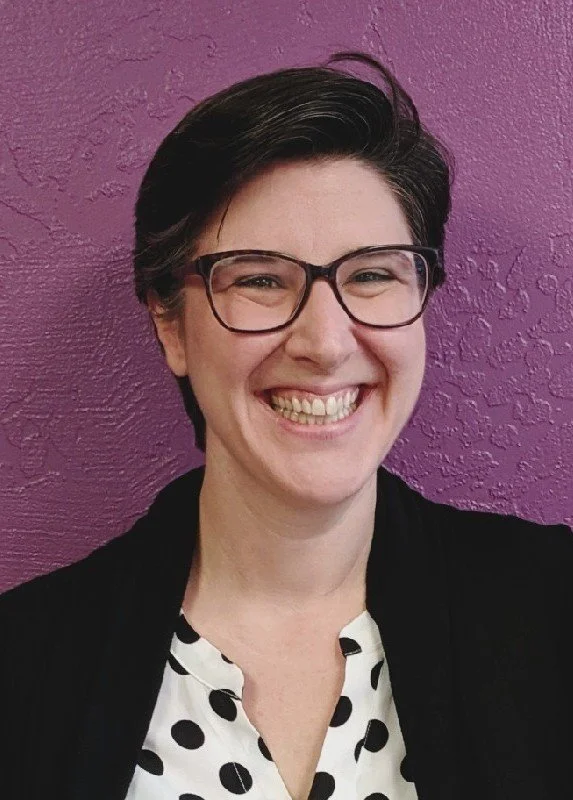 Smiling woman with short dark hair and glasses, wearing a black and white polka dot blouse, against a purple textured wall.