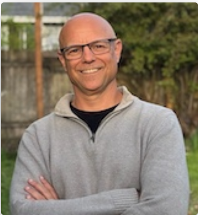 A smiling man with glasses, wearing a gray zip-up jacket, standing outdoors with a wooden fence and trees in the background.