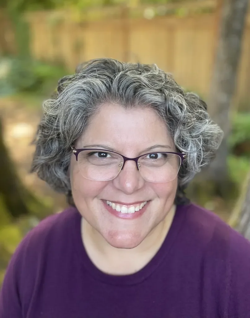 A smiling woman with curly gray hair and glasses, wearing a purple shirt, outdoors with blurred trees and a fence in the background.