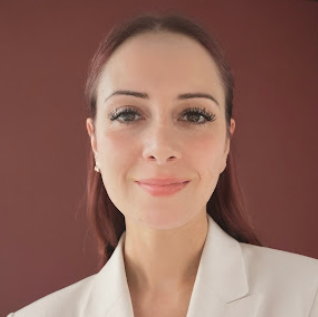 Close-up portrait of a woman with light makeup, auburn hair, and a white blazer, smiling softly against a brown background.