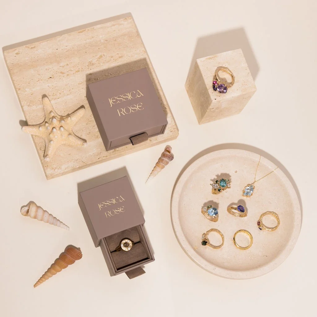 Jewelry display with rings, earrings, and necklaces, decorative seashells and starfish, and jewelry boxes labeled 'Jessica Rose' on a white surface.
