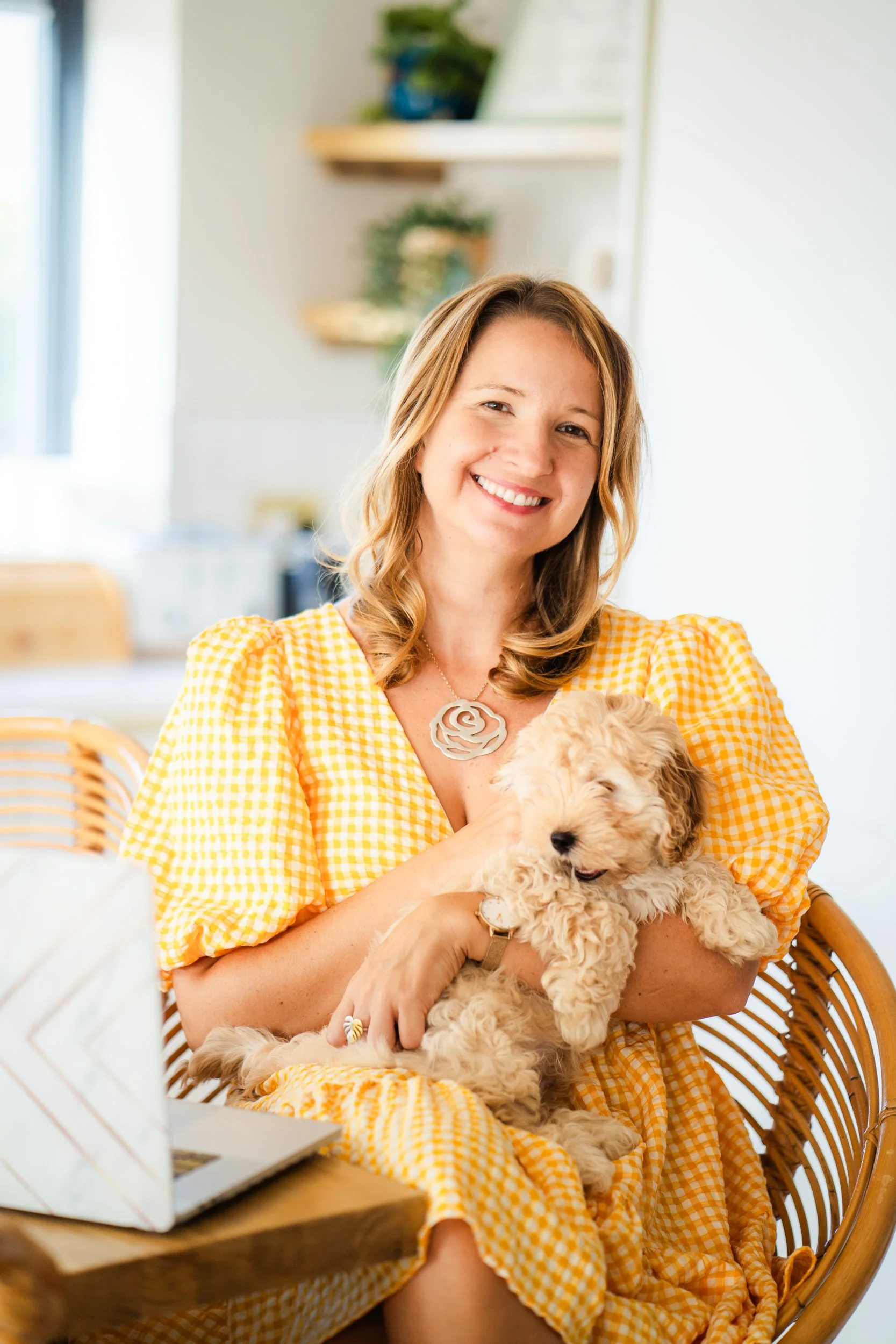 A smiling woman with shoulder-length light brown hair, wearing a yellow and white gingham dress and a large silver rose-shaped necklace, is sitting on a wicker chair holding a fluffy golden puppy in her arms in a bright, cozy room.