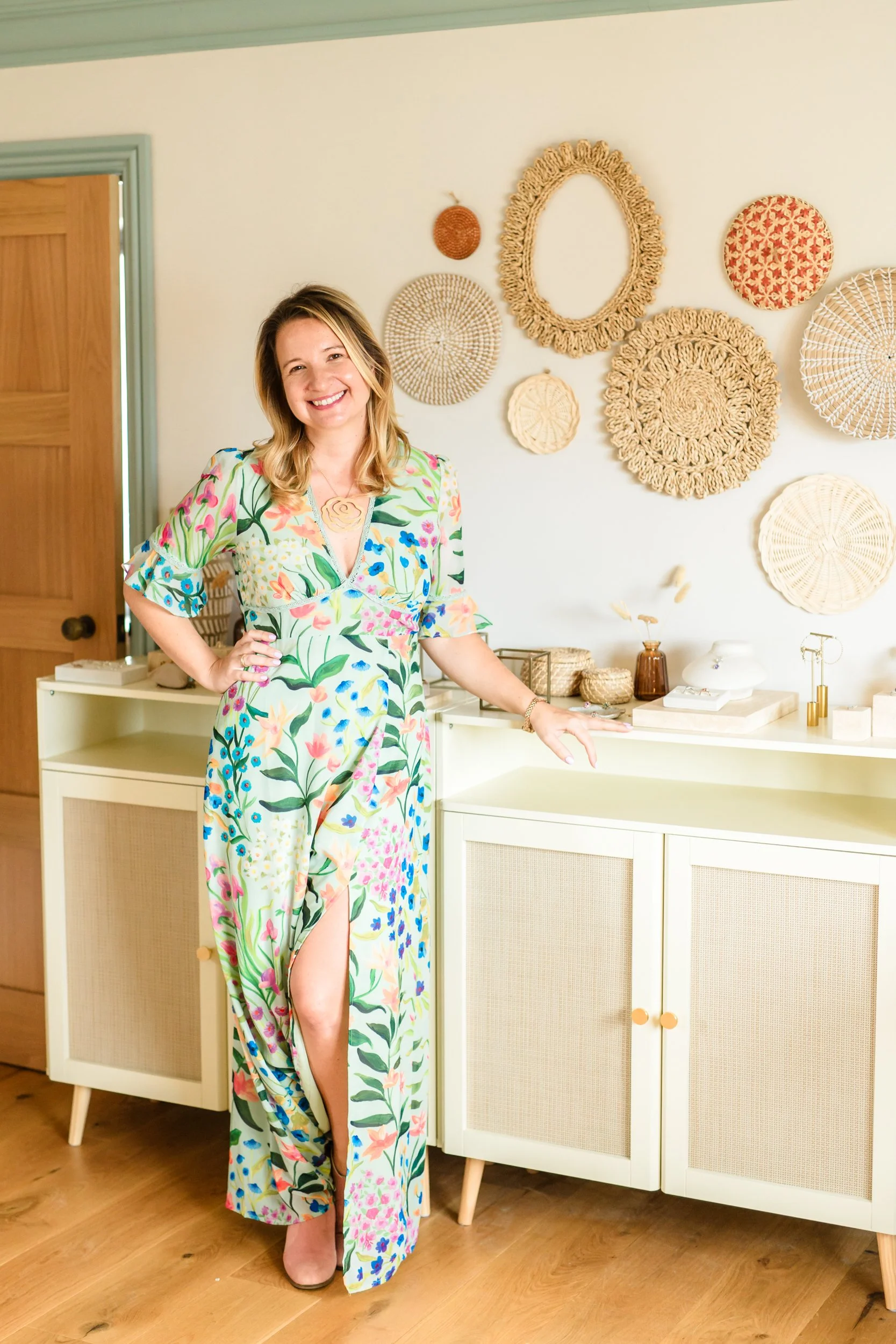 A smiling woman in a colorful floral dress standing indoors next to a white cabinet with decorative round woven wall hangings above it.