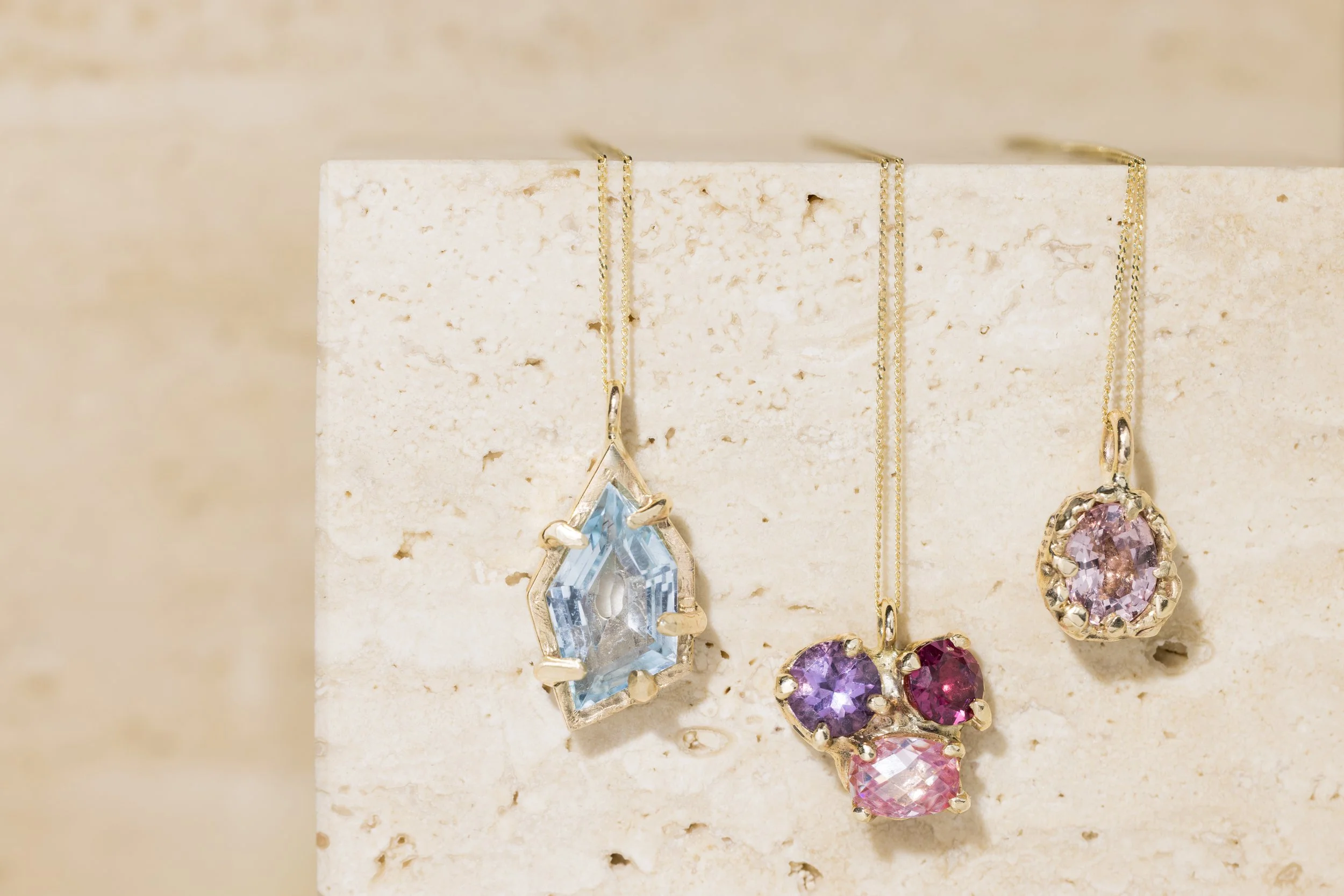 Three gold necklaces with large gemstone pendants hanging against a beige background.