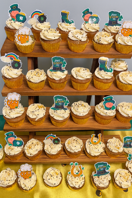 Stacked shelves displaying cupcakes decorated with leprechaun hats, rainbows, shamrocks, and beer mugs for St. Patrick's Day celebration.