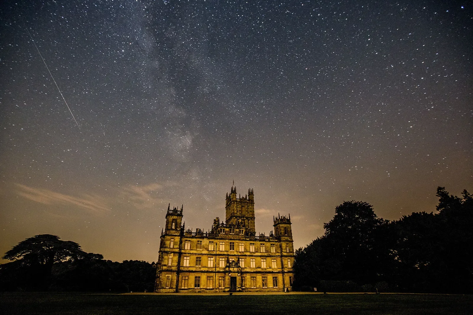 A large historic castle with towers and gothic architecture illuminated at night with a starry sky above.