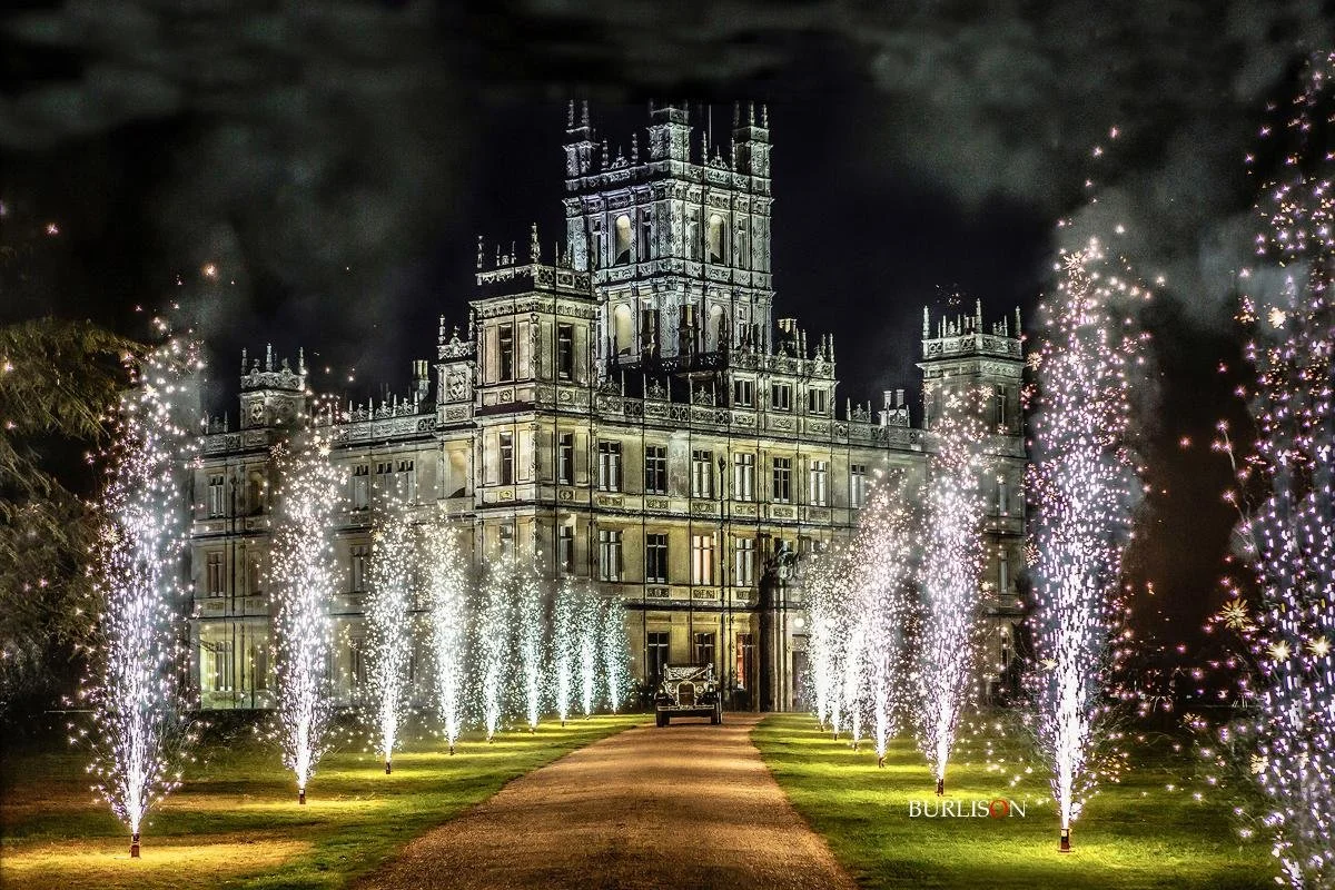 A large, ornate castle illuminated at night with fireworks shooting up along the driveway leading to the entrance.