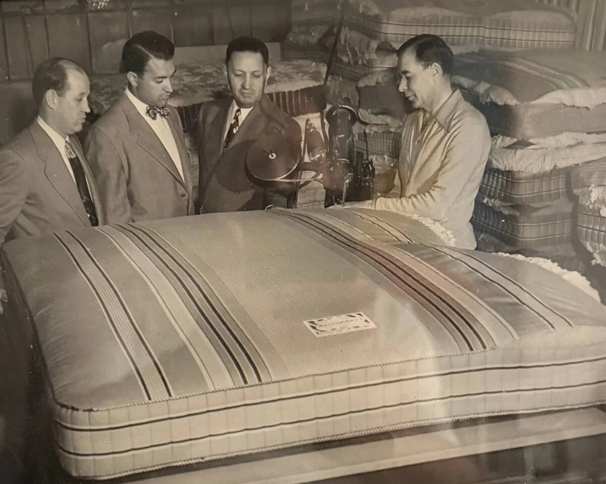 Four men in suits standing around a young man sitting at a sewing machine, in a room with mattresses stacked against the wall.