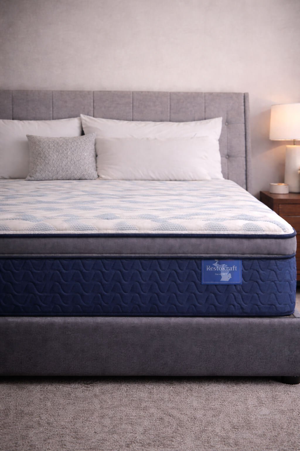 A neatly made bed with white and gray pillows, a light gray upholstered headboard, a blue mattress, and a wooden nightstand with a lamp on it.