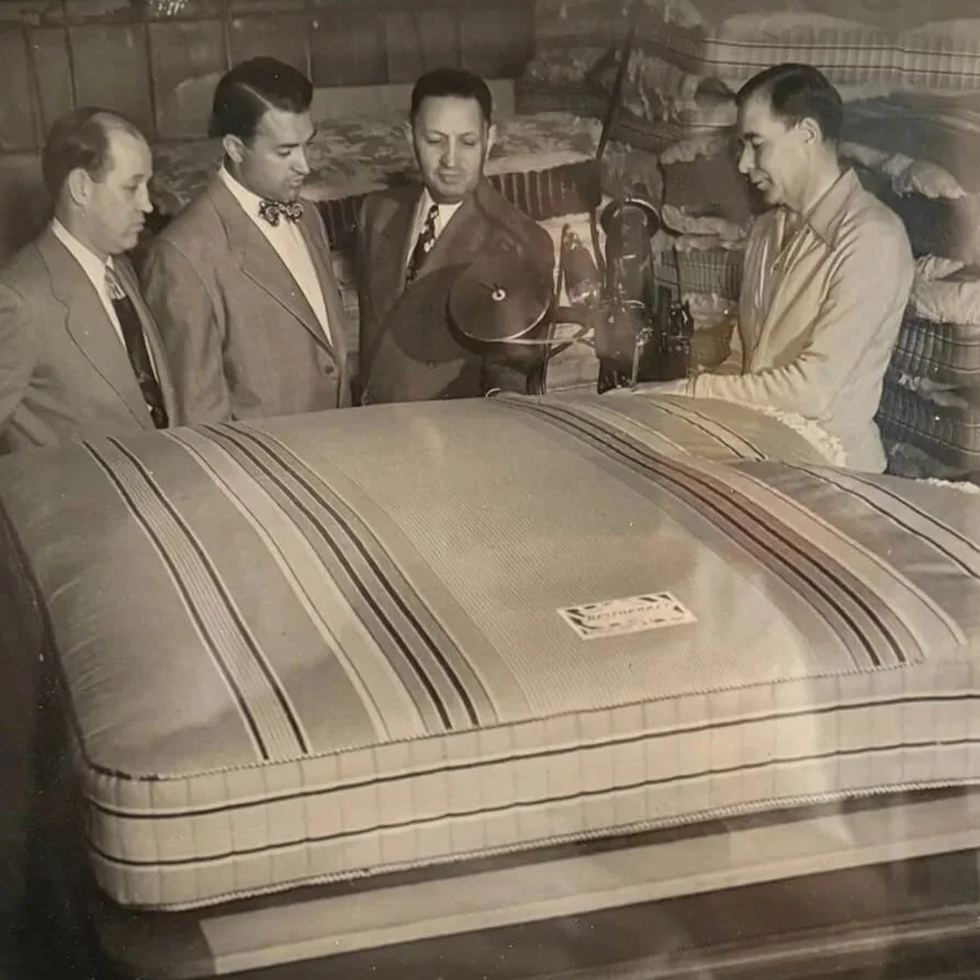 Four men in suits standing around a large covered mattress in a showroom or warehouse, with stacks of bedding and furniture in the background.
