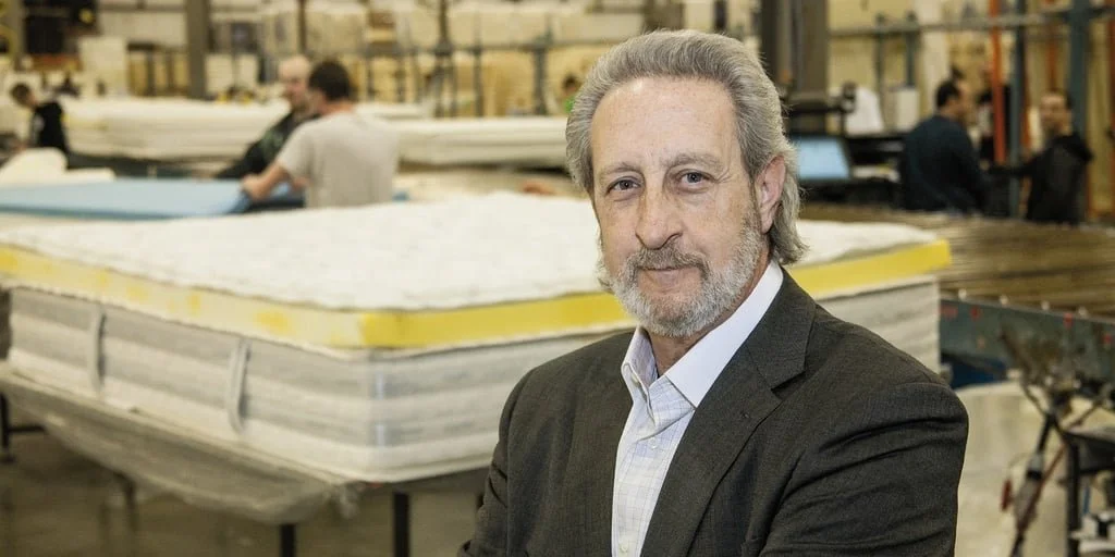 A middle-aged man with gray hair and a beard, wearing a suit, standing in a manufacturing or warehouse setting with industrial equipment and workers in the background.