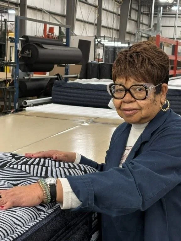 An elderly woman wearing glasses, a navy jacket, and a watch, inspecting fabric in a manufacturing warehouse.