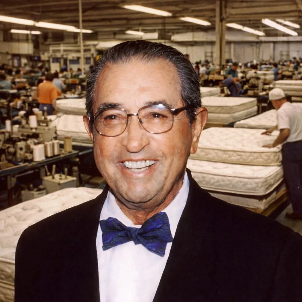 A man dressed in a tuxedo with a blue bow tie, smiling in an industrial mattress manufacturing plant.