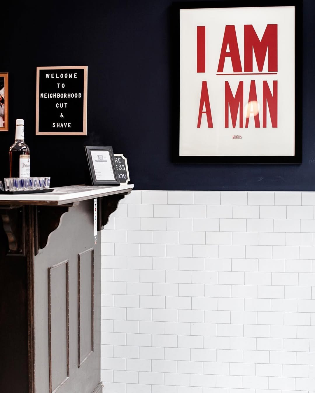 Indoor scene of a barbershop with framed signs on a dark wall, including a large white poster with red text "I AM A MAN" and a smaller black sign reading "WELCOME TO NEIGHBORHOOD CUT & SHAVE." A wooden counter off to the side has a framed menu, a bot