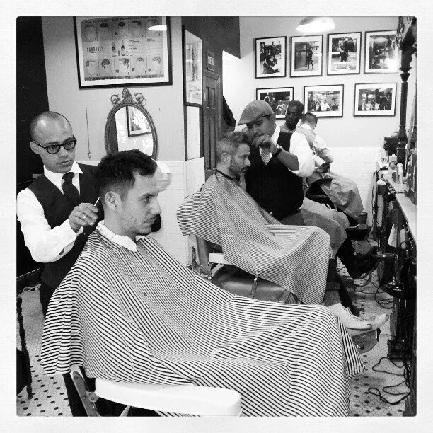Men getting haircuts at a barbershop with barber staff, framed photos on the wall, and vintage decor.