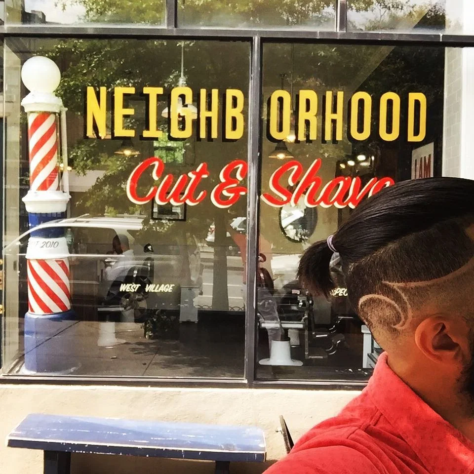 Barber shop window with yellow and red sign that reads 'Neighborhood Cut & Shave,' and a barber with a hairstyle featuring shaved designs on the side, sitting outside in a red shirt.