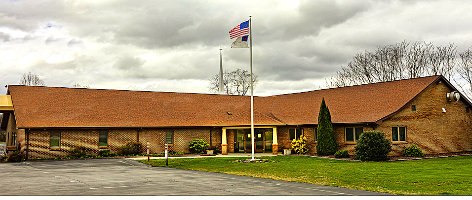 A brick building with a sloped reddish-brown roof and multiple windows. Two flags, the American flag and another flag, are flying on flagpoles in front. There is a parking lot and some greenery, including bushes and trees, under a cloudy sky.