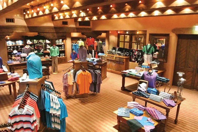 A golf pro shop featuring racks of colorful polo shirts, sweaters, and golf apparel. The store has wooden fixtures and warm lighting.
