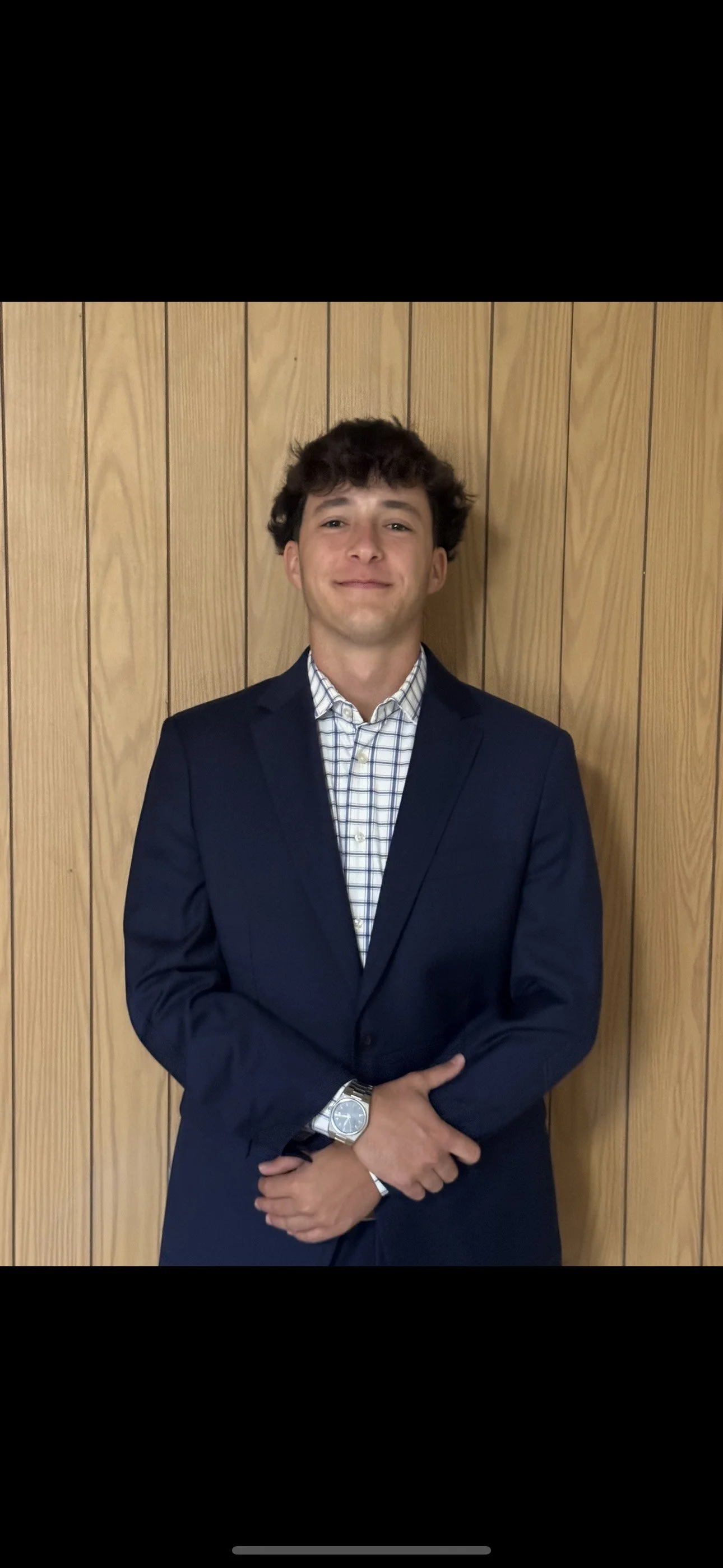 A young man in a dark blue suit with a checkered dress shirt stands against a wooden-paneled wall, smiling with arms crossed.