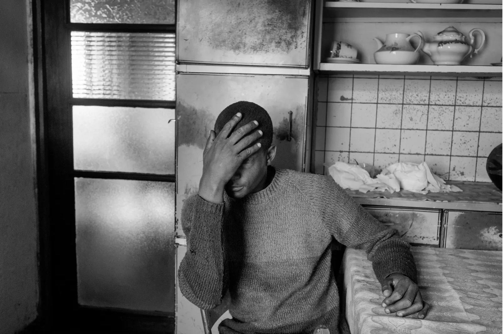 A young man with a hand on his forehead, sitting at a table in a kitchen with patterned tablecloths and open cabinets with dishes, in a black and white photo.