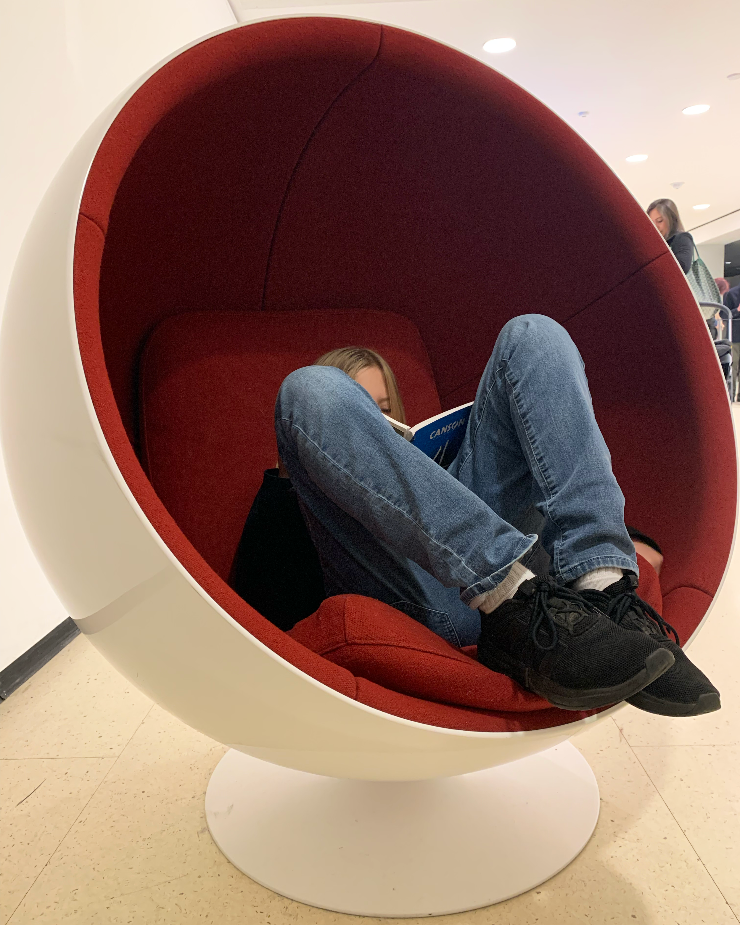 Person sitting inside a modern, circular chair with red upholstery, reading a book in a public space.