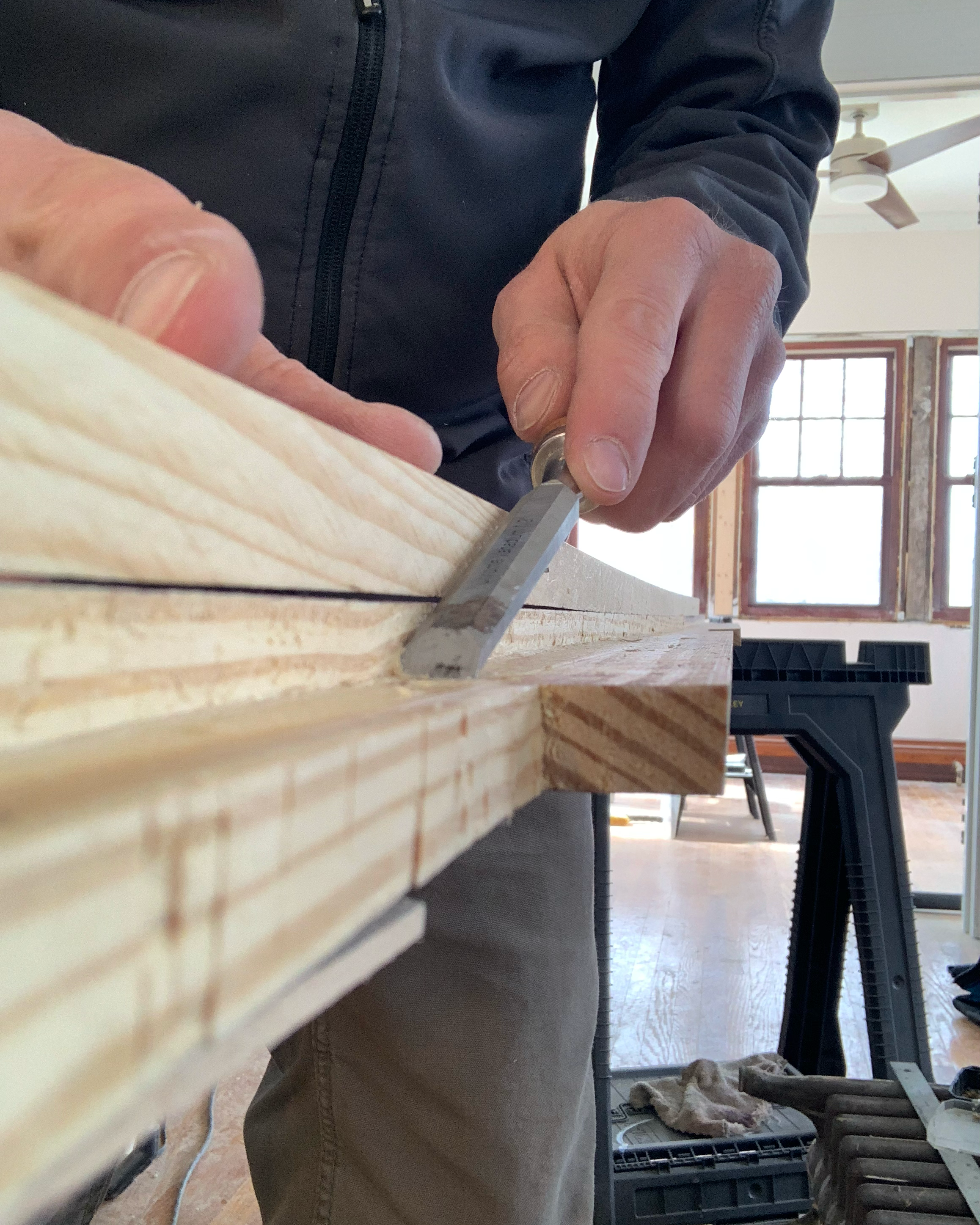 Person using a hand saw to cut a wooden board in a workshop with large windows and a ceiling fan.