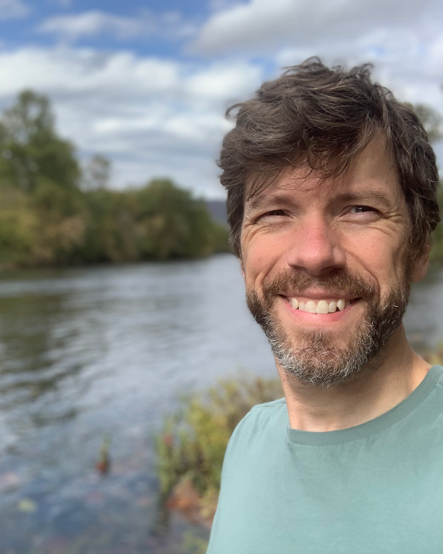 A smiling man with brown hair and a beard taking a selfie outdoors by a river, with trees and a cloudy sky in the background.