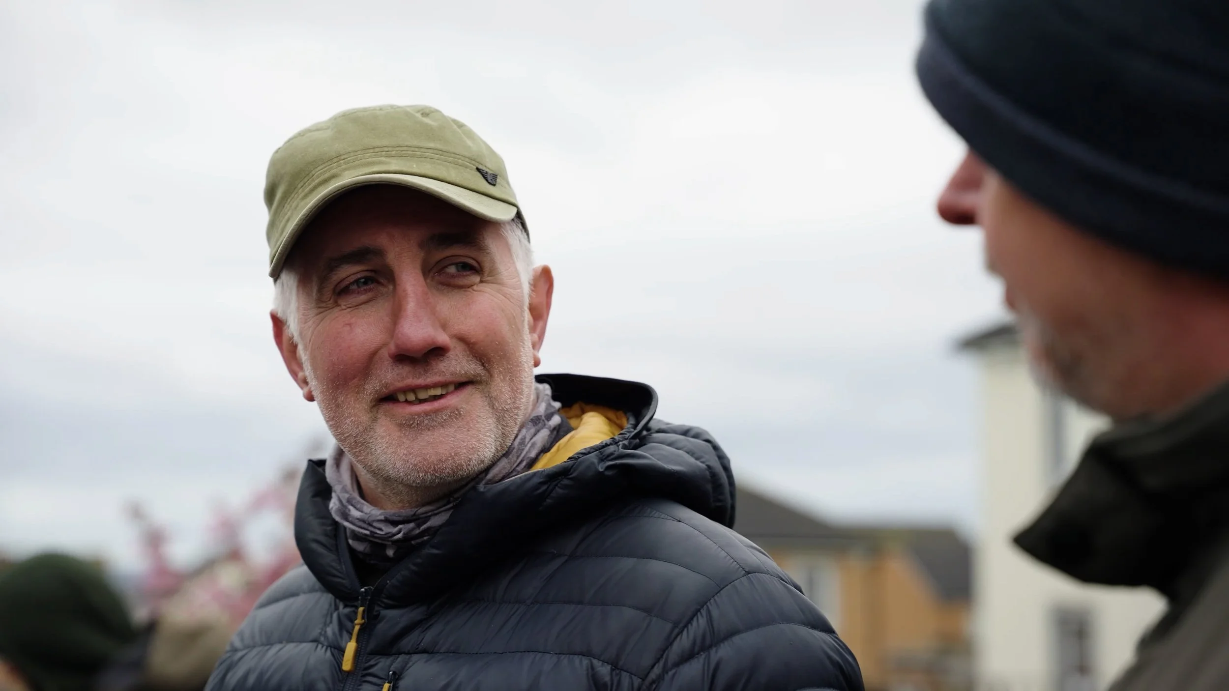 Two men outdoors, one smiling wearing a tan cap and jacket, the other partially visible in dark clothing and a black beanie, having a conversation on cloudy day.
