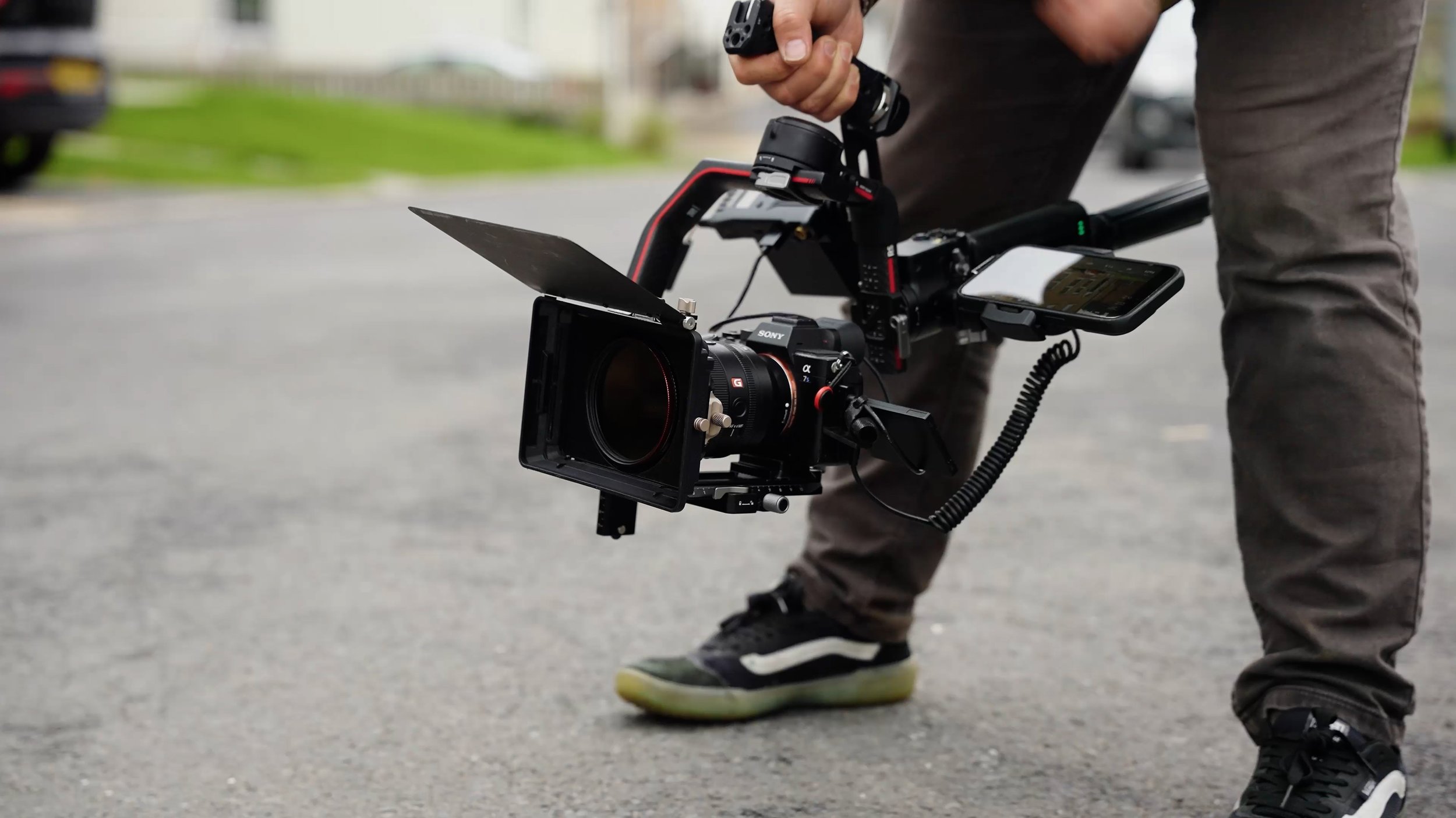 A person is adjusting a professional video camera mounted on a stabilizer rig outdoors, with a smartphone attached nearby, and wearing sneakers and casual pants.