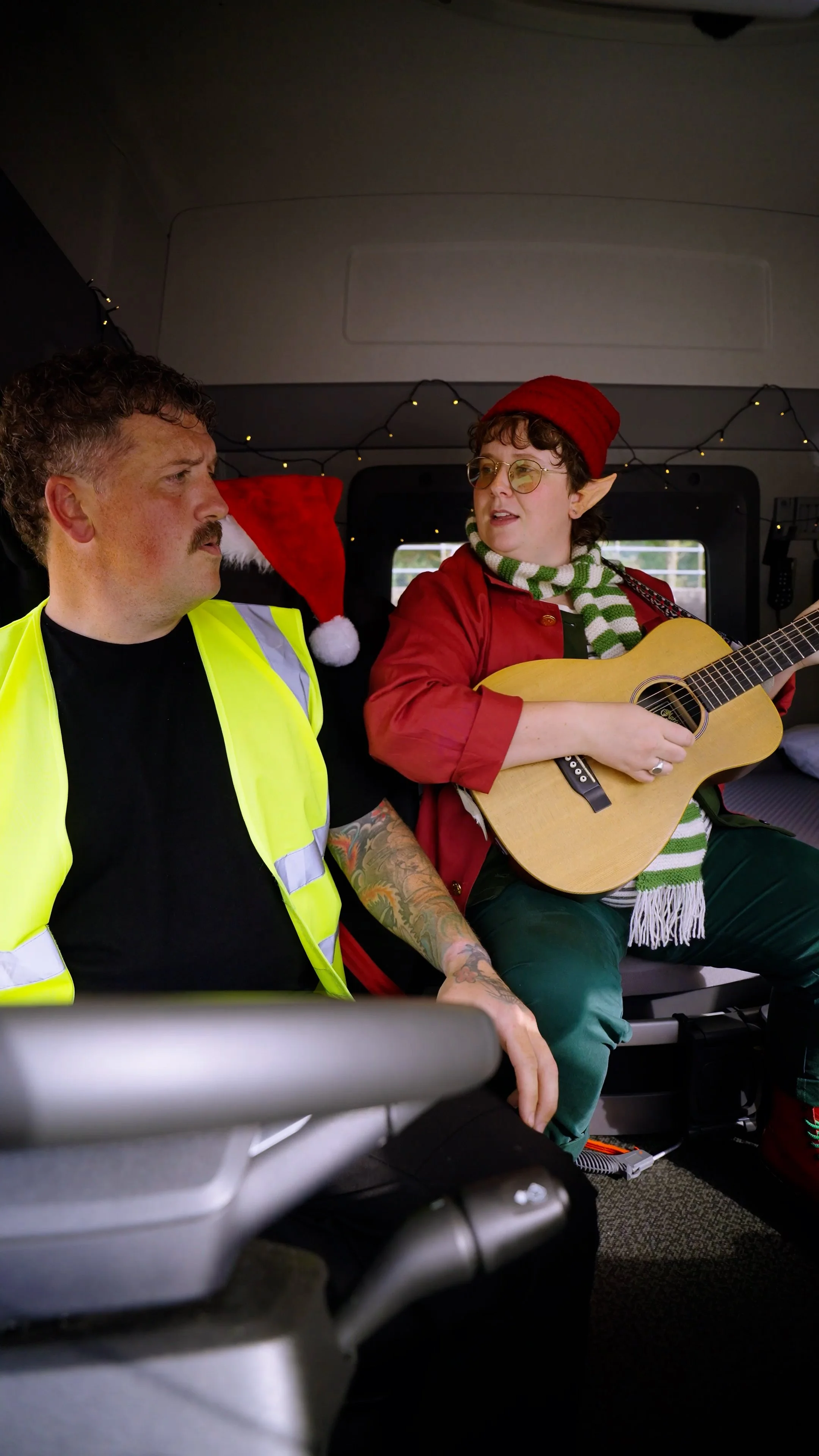 A man with a mustache and tattoos wearing a yellow safety vest sits next to a person dressed as an elf with elf ears, red hat, red jacket, and striped scarf, playing a guitar inside a vehicle decorated with Christmas lights.