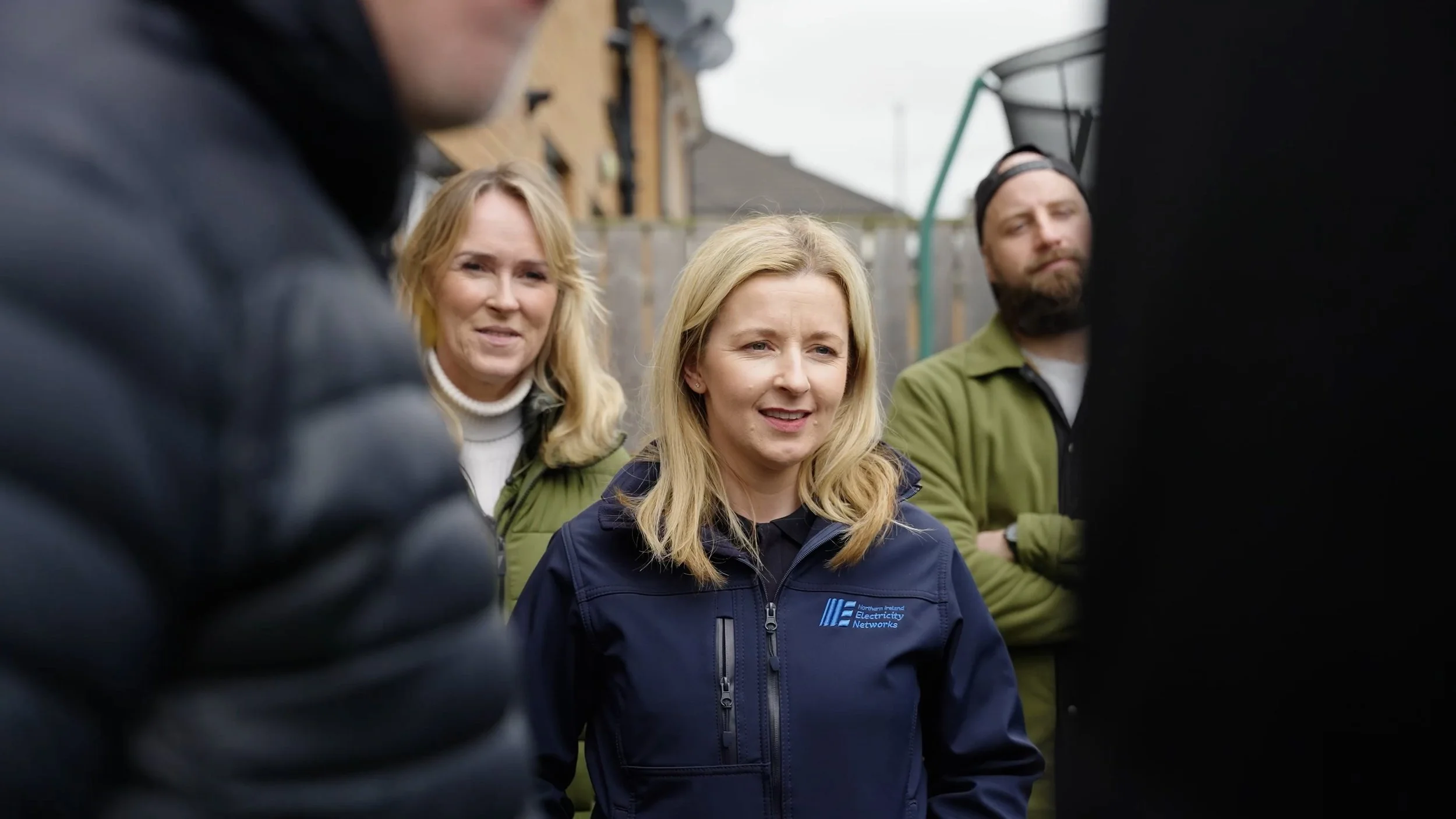 Group of four people outdoors, engaged in conversation, with one woman in a navy jacket bearing a logo, others in casual jackets, in a residential area with fencing and houses in the background.