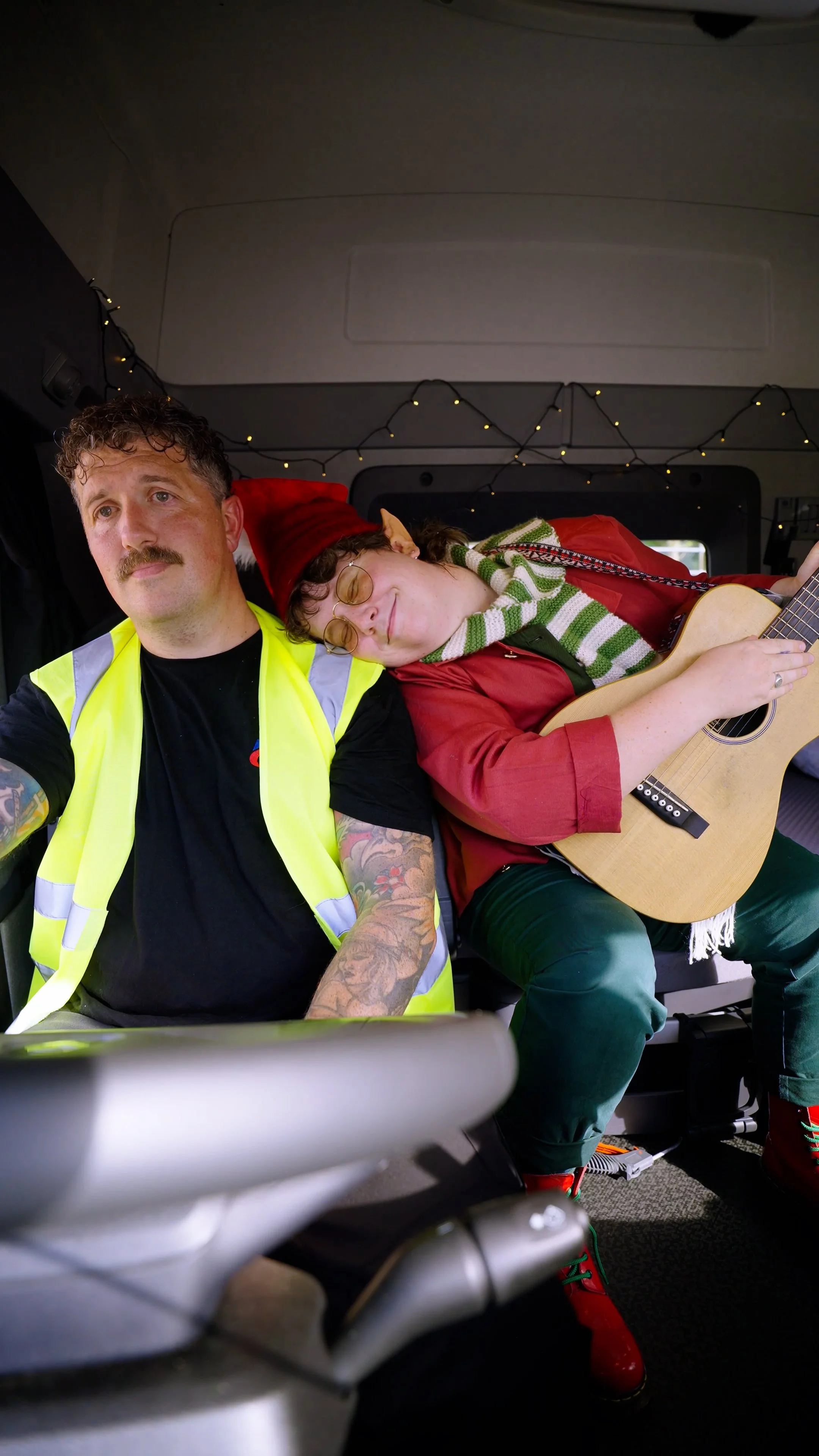 Two men sitting inside a vehicle, one wearing a yellow safety vest and the other in Christmas attire with a red elf hat, red jacket, and playing a guitar, while string lights decorate the space.
