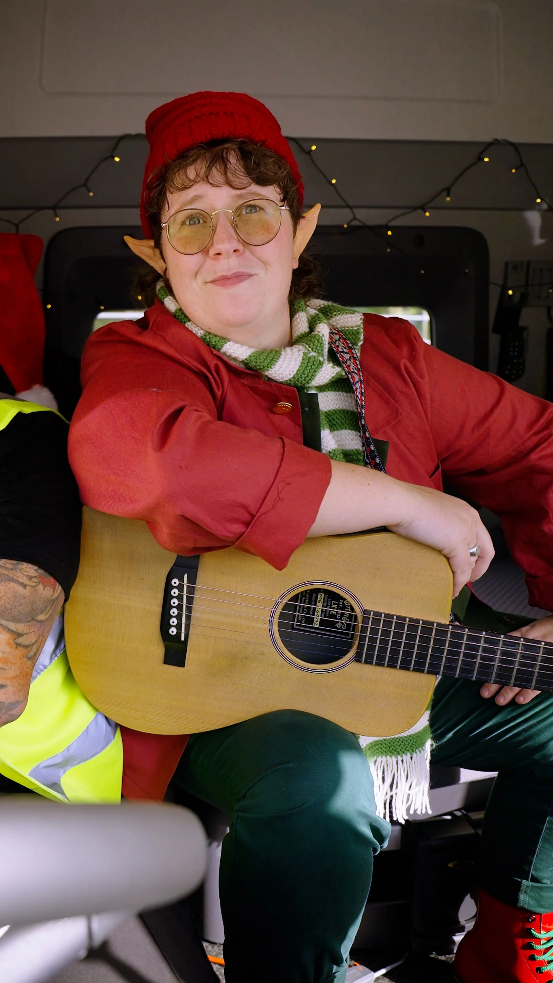 A person dressed as an elf with pointed ears, red hat, glasses, and a green and white striped scarf, holding a guitar, sitting inside what appears to be a vehicle decorated with holiday lights.