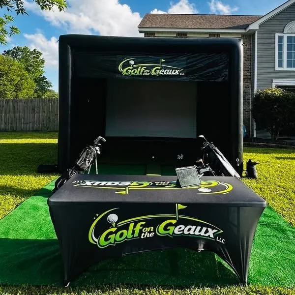 Portable golf simulator setup with a large screen, two golf clubs, and a tablet, branded "Golf on the Geaux LLC," on a green carpet outdoors.